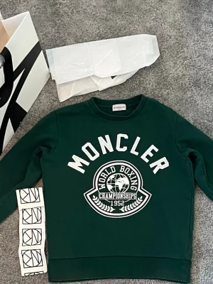 Moncler sweatshirt  - Mörkgrön sweatshirt från Moncler med vit text och tryck framtill. Tröjan har rund halsringning och ribbade muddar vid ärmslut och nederkant. Alla tillbehör ingår och pris kan självklart diskuteras