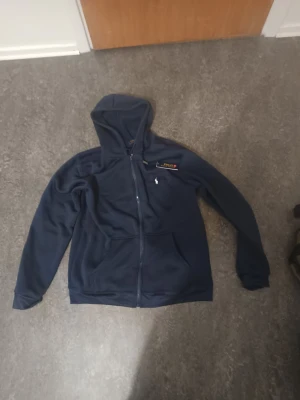 Mörkblå hoodie Polo Ralph Lauren - Snygg mörkblå hoodie Polo Ralph Lauren med dragkedja framtill och klassisk huva. Ikoniska Polo-loggan broderad på bröstet. Mjukt material och två fickor framtill. Perfekt för en avslappnad och stilren look.