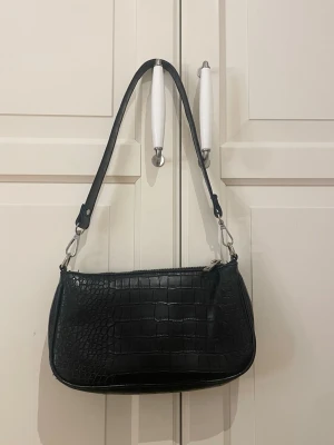 Svart axelväska i croco-mönster - Snygg svart handväska med croco-mönstrad yta och silverfärgade detaljer. Väskan har en avlång, mjukt rundad form och ett justerbart axelband. Perfekt för dig som gillar trendiga accessoarer med lite edge.
