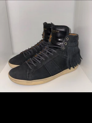 Saint Laurent svarta höga sneakers mocka - Svarta höga sneakers från Saint Laurent storlek 42 i mocka med fransdetaljer på hälen och svart läder vid plösen. Klassisk gummisula i beige och matchande svarta skosnören. Snygg och unik design som sticker ut.