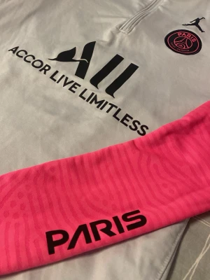 PSG x Jordan träningströja grå/rosa - Snygg träningströja från PSG x Jordan i ljusgrått med rosa ärmar och svarta detaljer. Tröjan har halv dragkedja, broderad Jumpman-logga och PSG-emblem på bröstet samt tryckt sponsorlogga. Perfekt för fotbollsträning eller streetwear. Jätte fina mönster på armen. Absolut inga fel på den. Använd ett par gånger. Går att ha på träningen men även vid annan tillfällen. Står på den 158-170 cm men tror den hade även passat för xs/s.