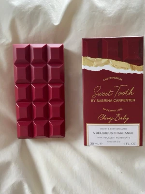 Sweet Tooth Cherry Baby parfym - Parfymen Sweet Tooth Cherry Baby från Sabrina Carpenter kommer i en unik, mörkröd flaska formad som en chokladkaka. Doften är söt och sofistikerad, och flaskan rymmer 30 ml. Förpackningen är också mörkröd med gulddetaljer och elegant design. Bara ett fåtal sprut tagna 