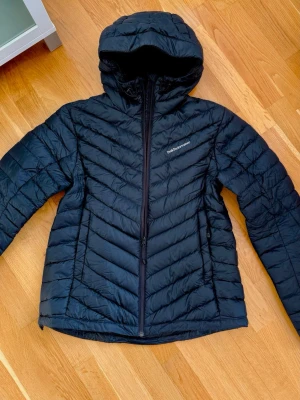Peak Performance Frost Svart dunjacka i mycket fint skick - Peak Performance Frost Down Hood jacka i storlek L. Klassisk modell som är både varm, bekväm och lätt att bära. Jackan fungerar lika bra som ytterlager som under en skaljacka för extra värme. Är fylld med 90/10-ankdun/fjädrar av hög kvalitet för exceptionell värme. Elastiska ärmslut, fickor med dragkedja och en tättslutande huva som ger maximalt skydd. Innerficka med dragkedja. Perfekt för kalla vinterdagar men fungerar även bra under vår och höst. Endast använd ett par gånger.
