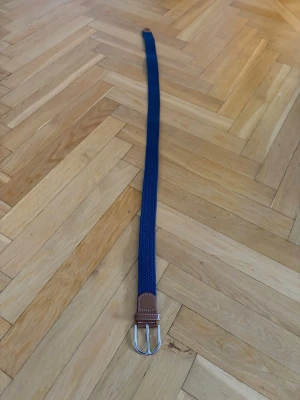 Blått flätat bälte  - Snyggt blått flätat bälte med bruna skinn-detaljer vid spännet. Bältet har ett klassiskt silverfärgat spänne och passar perfekt till jeans eller chinos. Enkel och stilren design som lyfter din outfit.