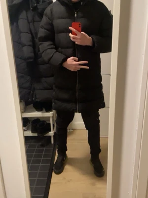 Lång svart dunjacka med huva - Säljer en lång svart dunjacka med huva och dragkedja framtill. Jackan har en clean look och puffig design som håller dig varm under kalla dagar. Perfekt för dig som vill ha en stilren vinterjacka med extra längd.