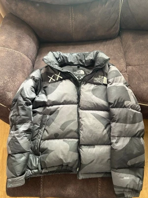 Svart camo dunjacka The North Face - Säljer en svart och grå camo dunjacka från The North Face x KAWS med 700-fill power. Jackan har dragkedja, hög krage och ikoniska korsdetaljer på bröstet. Perfekt för kalla vinterdagar och riktigt snygg streetwear.Zip handen är sönder men det går fortfarande att zippa upp och ner utan problem. En riktigt fin jacka en fynd. Jag är öppen för prisförslag jackan kostar runt 400$ men jag säljer min för 80$