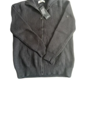 Svart stickad ziptröja Stone Island - Svart stickad ziptröja från Stone Island med klassisk logotyp på ärmen. Tröjan har hög krage, ribbstickad struktur och dragkedja framtill. Perfekt för dig som gillar mode och vill ha en stilren look.