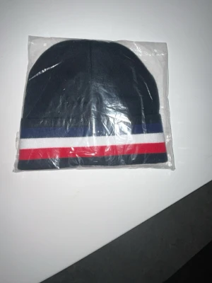 Mörkblå Moncler mössa med ränder - Snygg mörkblå mössa från Moncler med klassiska ränder i blått, vitt och rött längs kanten. Mössan har Moncler-logga framtill och är tillverkad i mjukt stickat material. Perfekt accessoar för att hålla stilen när det är kallt ute. Pris kan diskuteras 
