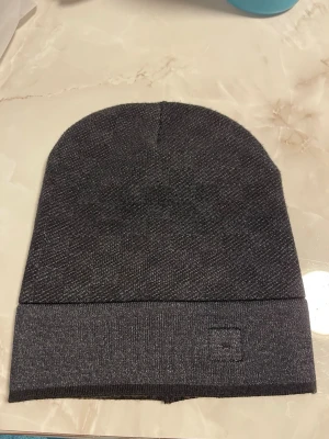 Mörkgrå ribbad mössa - Snygg mörkgrå mössa med uppvikt kant och diskret broderad logga framtill. Tillverkad i mjukt, ribbstickat material som sitter skönt på huvudet. Perfekt accessoar för kyliga dagar och enkel att matcha med olika outfits.