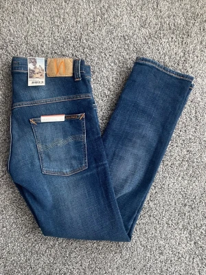 Blå jeans från Nudie Jeans - Snygga blå jeans från Nudie Jeans med klassisk femficksdesign och raka ben. Jeansen har en djupblå tvätt med subtila slitningar och kontrastsömmar. Tillverkade i bomull med Nudie-logga på bakfickan och läderpatch i midjan.