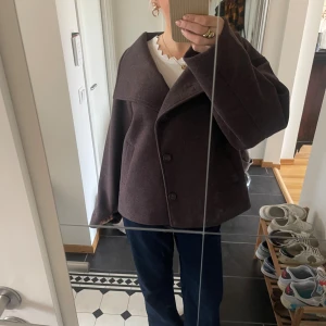 Brun ullkappa med bred krage - (Prislappen sitter kvar aldrig andvänds) Säljer en brun kappa i ull med bred krage och stora knappar framtill. Jackan har en loose passform, raka ärmar och två fickor på sidorna. Perfekt för höst och vinter, enkel att styla till jeans eller kjol. 