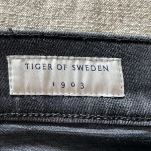 Mörkgrå jeans från Tiger of Sweden W28 L32 - Snygga mörkgrå jeans från Tiger of Sweden med klassisk femficksdesign och läderpatch bak i midjan. Jeansen har raka ben och är tillverkade i mjukt denimtyg som sitter skönt. Perfekta för dig som gillar stilrena och tidlösa plagg. Storlek 28/32