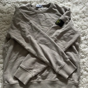 Beige/grön/grå aktig sweatshirt från Stone Island - Snygg beige ish sweatshirt från Stone Island med klassisk rund hals och ribbade muddar. Tröjan har den ikoniska Stone Island-patchen på vänster ärm och är tillverkad i mjuk bomull. Perfekt för dig som gillar stilrena och trendiga plagg. Den är storlek 156 passar mej som använder xs/s. Detta är mer av en intressekoll om någon skulle vara intresserad är det bara skriva!!
