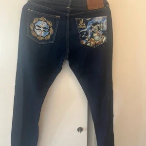 Evisu jeans med broderade bakfickor - Mörkblå Evisu jeans med unika broderier på bakfickorna – ena fickan har ett japanskt motiv med texten 'FUJIN' och den andra har ett stiliserat mönster i blått och guld. Klassisk femficksmodell i jeansmaterial med raka ben och snygga kontrastsömmar. Köpt för 3600kr i NK Stockholm år 2022! Använda ett fåtal gånger så dem är i bra skick! Säljer för jag inte använt dem!