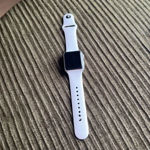 Apple Watch med vitt armband - Apple Watch, (serie 3) med fyrkantig svart display och vitt silikonarmband. Klockan har silverfärgad boett och kommer med original laddare. Perfekt för dig som vill ha koll på notiser, träning och hälsa direkt på handleden.