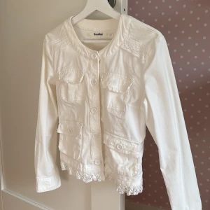 Vit jacka med spets från RoseBud - Vit jacka från RoseBud med snygga spetsdetaljer på axlar, längs knäppningen och nederkanten. Jackan har fyra fickor framtill, stora vita knappar och lång ärm. Perfekt för dig som gillar en söt och stilren look med extra detaljer.