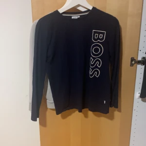 Mörkblå långärmad tröja från BOSS - Snygg mörkblå långärmad tröja från BOSS med stort vitt logotryck längs ena sidan framtill. Tröjan har rund halsringning och är tillverkad i mjuk bomull, perfekt för en clean och stilren look.