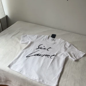 Vit Saint Laurent t-shirt med tryck - Säljer en clean vit t-shirt från Saint Laurent med svart handskrivet logotryck på bröstet. Klassisk rund hals och korta ärmar. Tillverkad i mjuk bomull för en skön känsla. Perfekt för dig som gillar stilrena designer och vill sticka ut med ett ikoniskt märke.