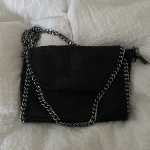 Svart axelväska med kedjedetaljer - Snygg svart axelväska i mocka med silverfärgad kedja som axelrem och runt kanterna. Väskan har lock med magnetknäppning och dragkedja på insidan. Perfekt storlek för det viktigaste och ger en cool vibe till din outfit.