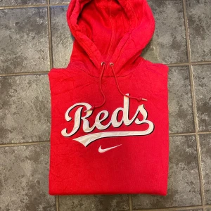 Röd Nike Reds hoodie med huva - Perfekt hoodie nu när hösten och vintern är här. Klarröd med stort Reds-tryck i vitt och svart på bröstet. Klassisk huva med snören och en stor magficka som håller händerna varma.  En färgstark och sportig hoodie som funkar både till vardags och när du vill sticka ut lite extra.
