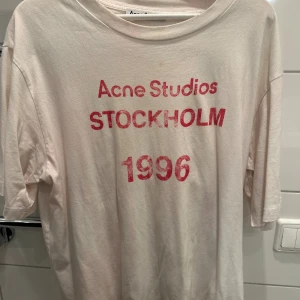 Vit Acne Studios t-shirt med tryck - Vit t-shirt från Acne Studios med rött tryck på bröstet där det står 'Acne Studios STOCKHOLM 1996'. Klassisk passform med rund hals och korta ärmar. Mjuk och skön i bomull, perfekt för en avslappnad stil.
