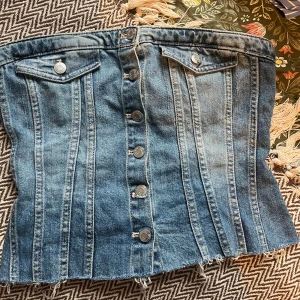 Jeans tubtopp från Zara med knappar - Säljer en snygg jeans tubtopp från Zara i medium. Toppen har knappar framtill, två bröstfickor med lock och rå kant nertill. Baksidan har smock för extra stretch och passform. Perfekt för dig som gillar denim och vill sticka ut.