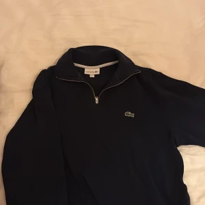 Mörkblå half zip tröja från Lacoste - Snygg mörkblå tröja från Lacoste med klassisk krokodil-logga på bröstet. Tröjan har en halv dragkedja och hög krage, perfekt för lager-på-lager. Tillverkad i mjuk bomull som känns skön mot huden. Enkel och stilren design som funkar till det mesta.