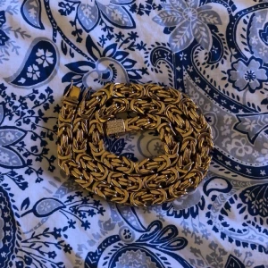 18K guldplatterad guldkedja med stenar - Säljer en kedja i 18K plattering guld med flätad kedja och lås dekorerat med gnistrande stenar. Kedjan har en riktigt maffig känsla och glänser snyggt. Perfekt för dig som vill sticka ut med ett statement-smycke. 70cm lång och 1cm bred 