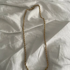 Guldfärgat halsband  - Säljer ett snyggt guldfärgat halsband. Kedjan har en tvinnad design som ger en cool och trendig look. Perfekt accessoar för att lyfta din outfit. Levereras i original svart sammetsfodral med guldtext. Platerad guld 18 karat, 50 cm 3mm bredd. 