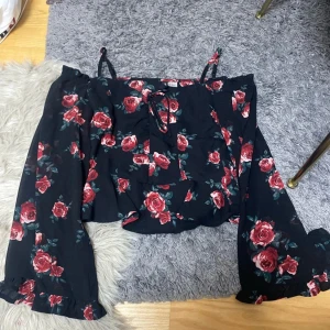 Blommig offshoulder topp från H&M Divided - Supersöt svart offshoulder topp med röda rosor från H&M Divided. Toppen har långa vida ärmar, smala axelband och en liten knytning framtill. Perfekt för dig som gillar romantiska vibes och blommigt mönster.