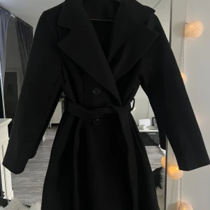 Svart kappa med bälte och knappar - Säljer en svart kappa med klassisk krage, knappar framtill och matchande bälte i midjan. Jackan har lång ärm och en stilren, tidlös design som passar perfekt till kyligare dagar. Materialet känns mjukt och värmande, perfekt för höst och vinter.