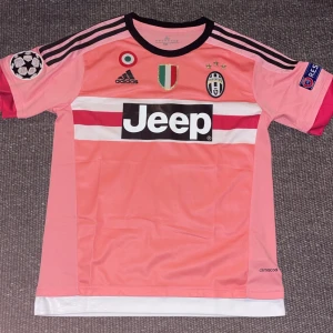 Juventus Dybala #21 rosa bortatröja - Juventus bortatröja i rosa med svarta och vita detaljer, Adidas-logga, Jeep-sponsor och Dybala #21 på ryggen. Kortärmad modell i lätt och ventilerande climacool-material. Champions League-patch på ärmen och klubbmärke på bröstet. Helt oanvänd