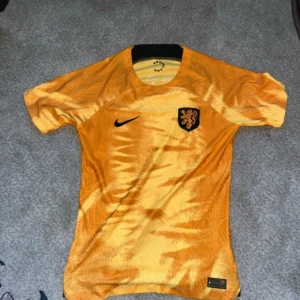 Holland fotbollströja Nike orange - Säljer en orange Nederländerna fotbollströja från Nike med svart lejonmärke på bröstet och svart Nike-logga. Tröjan har korta ärmar, rund hals och är tillverkad i ett lätt, ventilerande material. Snyggt mönster i olika orange toner som ger en unik look.