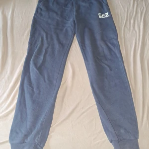 Blå mjukisbyxor från EA7 Emporio Armani - Mörkblå mjukisbyxor från EA7 Emporio Armani med logga på vänster höft. Byxorna har resår och snörning i midjan samt muddar vid bensluten. Tillverkade i mjukt material som passar perfekt för chill eller träning.