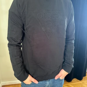 Svart långärmad tröja från Guess - Svart långärmad tröja från Guess med det klassiska broderade logotyptrycket framtill. Tröjan har rund halsringning och ribbade muddar vid ärmslut och nederkant. Tillverkad i mjuk bomull som är skön att bära hela dagen.