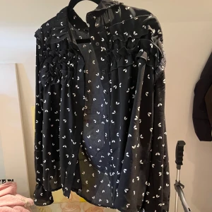 Svart blus med hjärtan från Chica London - Svart blus från Chica London med vita hjärtan, volangdetaljer över bröstet och knappar framtill. Långärmad och luftig modell som ger en cool och lekfull vibe. Perfekt för dig som gillar unika detaljer och mönster.