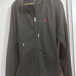 Svart Jordan hoodie med dragkedja - Svart hoodie från Jordan med röd broderad Jumpman-logga på bröstet. Tröjan har hel dragkedja, huva med snörning och två stora fickor framtill. Tillverkad i mjuk bomullsblandning och har långa ärmar. Perfekt för en sportig och avslappnad stil.