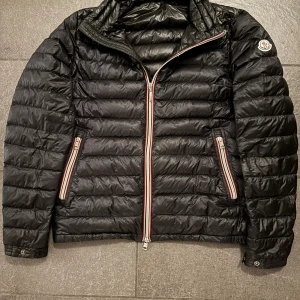  Moncler Daniel - Svart pufferjacka från Moncler, modell Daniel Giubbotto. Jackan har klassisk quiltad design, tvåvägsdragkedja och fickor med röd, vit och blå randiga detaljer. Moncler-logga på ärmen. Perfekt för kyliga dagar och riktigt snygg streetstil. Den är äkta och en lite äldre modell så Scanen på märket funkar ej men inuti funkar och kommer rätt och är 100% äkta storlek 4 är M-L jag som är 185 sitter den lite liten på. Stå jätte små hål men är lagningar över som ej syns 