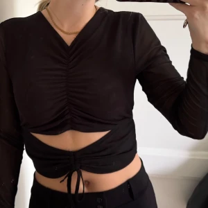 Svart croppad topp  - Svart långärmad croppad topp från FB Sister med v-ringning och snygg rynkdetalj framtill. Toppen har meshärmar och knytning nertill. Står storlek L men skulle säga att den är en S 