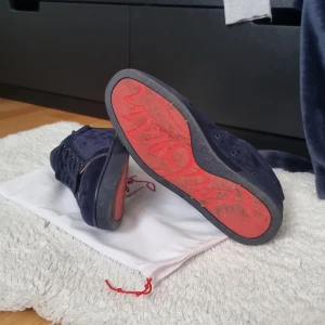 Mörkblå mocka sneakers från Louboutin - Snygga mörkblå sneakers från Christian Louboutin i lyxig mocka med klassisk röd sula och diskret logga undertill. Skorna har rund tå, platt sula och snörning framtill. Insidan är i ljust skinn och de kommer med original dustbag.