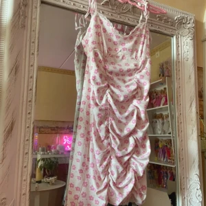 Dress No°2 - Barbiecore. Supersöt kortklänning med vita och rosa blommor, tunna axelband och volangig rynkning framtill. Klänningen har spetsdetaljer vid bysten och är gjord i ett mjukt, ribbat material. Perfekt för sommarens alla tillfällen!