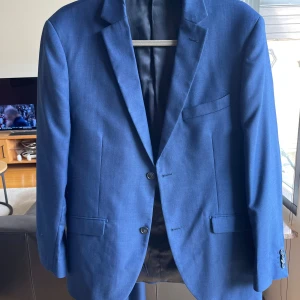 Byxornas storlek är 50 och kavajens är 52 - Snygg blå kostym från Riley Stanton med klassisk kavaj och matchande byxor. Kavajen har två knappar, smala slag och flera fickor. Kostymen är helfärgad och har en stilren, modern passform. Perfekt för dig som vill ha en stilren och tidlös look.