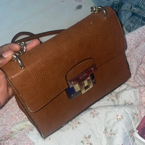 Brun axelväska från ALDO med kedja - Snygg brun axelväska från ALDO i skinnimitation med grov guldkedja och metallspänne framtill. Väskan har ett fack med dragkedja på baksidan och flera innerfack. Perfekt storlek för det viktigaste och ger en stilren vibe till din outfit.