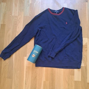 Marinblå tröja från Polo Ralph Lauren - Säljer en marinblå långärmad tröja från Polo Ralph Lauren med röd broderad logga på bröstet och röda detaljer vid halsringningen. Tröjan är i mjuk bomull och har klassisk passform med ribbade muddar vid ärmslut och nederkant. Nypris: 1049