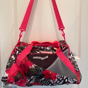 Färgstark träningsväska från Desigual - Säljer en unik träningsväska från Desigual med blommigt och grafiskt mönster i svart, vitt och rosa 🌸Väskan har både axelrem och handtag, flera fack med dragkedja och mesh-detaljer på sidorna. Perfekt för gymmet eller sportaktiviteter. Helt i nyskick, knappt använd 🌸