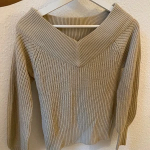 Beige stickad tröja med v-ringad krage - Beige stickad tröja med bred v-ringad krage som kan bäras offshoulder. Tröjan har grov ribbstickning och långa ärmar. Perfekt för dig som gillar en enkel men snygg stil med en twist.