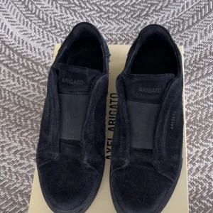 Svarta Axel Arigato skor - Svarta sneakers från Axel Arigato i mjuk mocka med elastisk panel över vristen istället för klassisk snörning. Snygg minimalistisk design med diskret logga på sidan och platt sula. Perfekta för dig som gillar stilrena och bekväma skor. Kartong och påse med kommer.