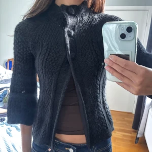 Svart stickad kofta från EastWest - Svart stickad kofta med snygg struktur och stora knappar framtill. Koftan har trekvartsärmar och hög krage, perfekt för lager på lager. I alpackamix. 