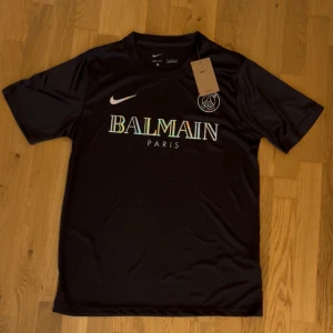 Svart PSG x Balmain fotbollströja Nike - Snygg svart fotbollströja från Nike i samarbete med PSG och Balmain. Tröjan har korta ärmar, rund hals och glansigt material. Framsidan pryds av Balmain Paris-logga i regnbågseffekt och PSG-emblem. Perfekt för dig som vill sticka ut på planen.