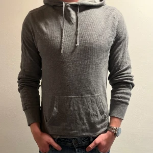 Grå hoodie från Polo Ralph Lauren - Snygg grå hoodie från Polo Ralph Lauren med diskret struktur i tyget. Tröjan har huva med snörning, magficka och en liten Polo RL-patch nertill. Perfekt för chill dagar och enkel att matcha med jeans eller joggers.